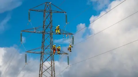 ENW View of the top part of an electricity pylon, with four two workmen on the lower stanchion, one on the middle one, and another on a ladder suspended between two wires. There is a patch of blue sky and somewhat ominous-looking white clouds.