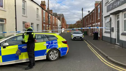 Blue and white police tape stretches from one side of a side street to the other. A street full of terraced houses can be seen with two police cars parked at the front of it. One male police officer with yellow high vis and an ear piece can be seen standing next to one of the cars.