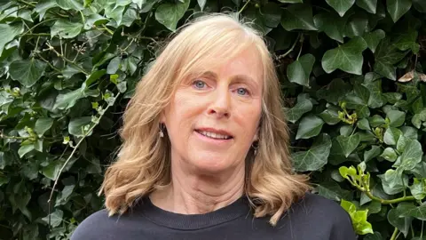 BBC/Marian McNamee Beverley has blonde hair that reaches her shoulders. She's wearing a dark blue top and standing in front of an ivy-filled hedge.