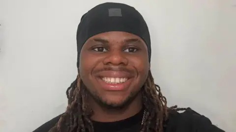 Tamani Clayton Tamani has brown and black dreadlocked hair and wears a cap, silver chain and black t-shirt. He has dark facial hair and smiles at the camera