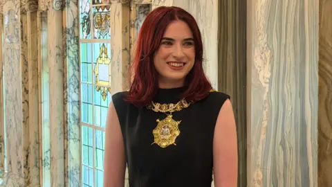 Róis-Máire Donnelly is smiling. She's wearing lipstick. She has red hair. She's wearing her gold chain of office and a black dress. A stained glass window is behind her and marble walls. 