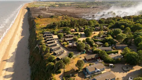 EPA/Shutterstock A bird's eye view of holiday homes, surrounded by trees, and near a cliff edge. Behind them is a smouldering black landscape. To the left is the sea.