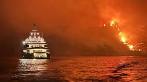 A photo of a luxury yacht on the water, with a raging fire on the land in the distance