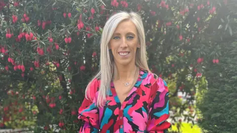 A woman is standing outside in front of a tree with pink buds. She is tall and has blond hair. She is wearing a bright dress which has various shades of pink, black and blue.