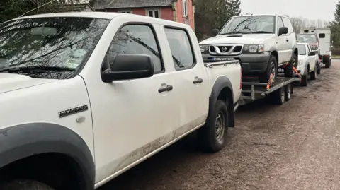 A convoy of white vehicles are lined up. A white pick-up truck is towing a trailer with a white SUV vehicle on it. 