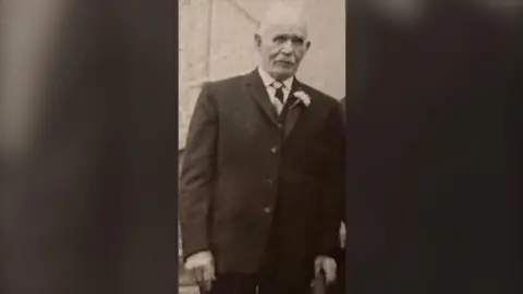 Family photo A black and white archive photo of the late Owen Pyers attending a wedding.  He is an elderly man with white hair.  He is wearing a dark suit, white shirt, a striped tie and has a flower in his buttonhole.