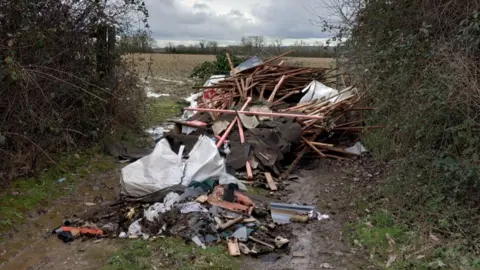 Wiltshire Council A large pile of debris made up of planks of wood, roofing felt and other items dumped in the gateway to a field. 