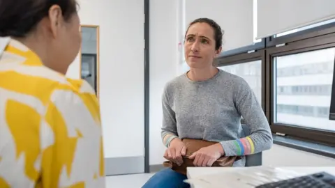 Getty Images An over the shoulder view of a female GP speaking to her male patient who has a concerned look on her face. She is seeking advice and how to deal with her ailment.