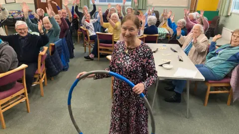 Justin Dealey/BBC Nova Strange holding a hula hoop while an audience cheers behind her, with their hands in the air.