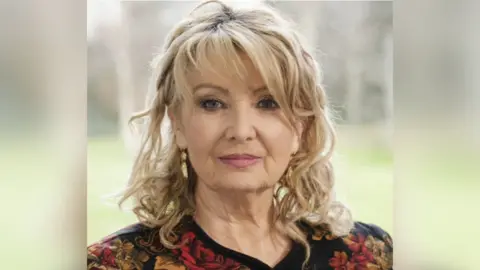 Marie Keenan An image of Prof Marie Keenan looking at the camera with a neutral expression on her face. She has short, curled blonde hair and is wearing a black, red and yellow patterned shirt. She is standing outside, but the background is blurred.