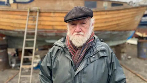 Ian Clark - man with white beard, wearing a grey jacket and multicoloured scarf and black hat standing in front of a small wooden fishing boat with a ladder perched next to it