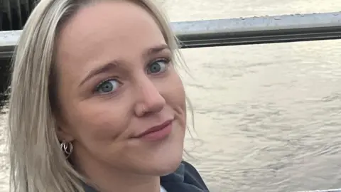 Chloe Wilson stands in front of a river. She has long blond hair and blue eyes, her face is close to the camera and she is wearing a nose ring and earrings. The collar of her grey jacket can also be seen