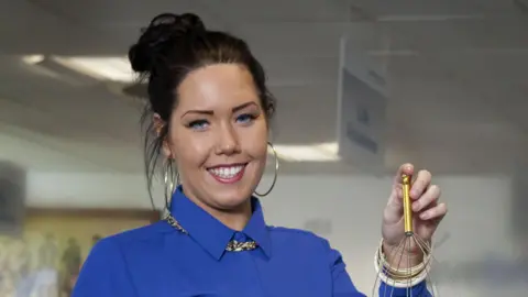 Hayley when she was in The Call Centre. She is wearing her hair up in a bun, is wearing a blue shirt and has large hoop earrings. 