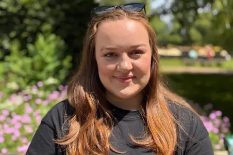 BBC Wales A woman in her late twenties with long hair, wearing a black top, smiling at the camera. She's sat in a public park with greenery behind her.