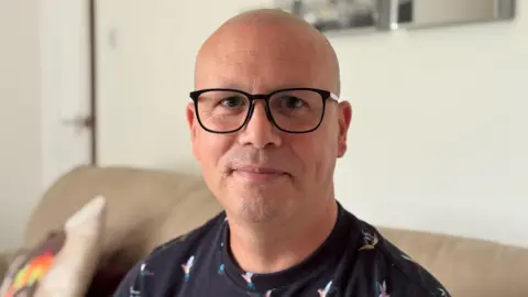 From a white-walled front room Jason Pye, with a shaven head, black-rimmed glassed and wearing a navy blue patterned top, looks into the camera lens from his beige leather sofa.