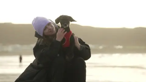 A young girl sat on the beach with a purple hat and black coat on, smiling at her black and white dog who is sat on her knees wearing a red harness.