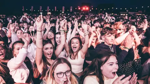 Hundreds of young people jump and wave and yell as they face towards a stage during the night.