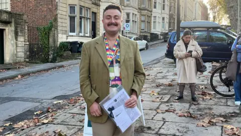 Ben Wilkinson-Turnbull Ben wearing a brown blazer and colourful lanyard standing on a street holding a folder, smiling at the camera.