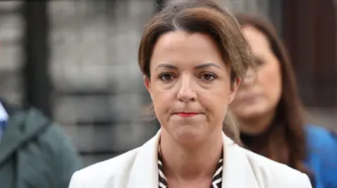 PA Media Infrastructure Minister Liz Kimmins speaking to the media outside Belfast High Court - she has dark hair and is wearing a white blazer and black and white top.