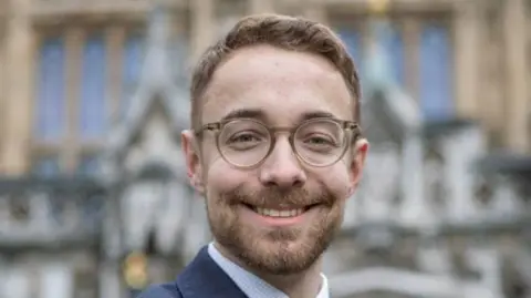 House of Commons A man pictured in front of the House of Commons. He is smiling, has brown framed glasses and a brown beard