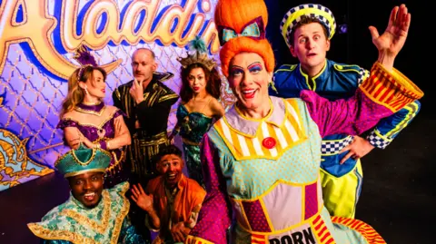 Shropshire Council Seven people, men and women, are dressed up in colourful costumes, with two of them particularly prominent in the image on the right. The word Aladdin is in large yellow letters on a mainly purple background behind them.