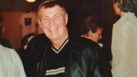 Family photograph Leslie Cawood pictured at a social event. He has short dark hair and is wearing a black leather jacket, V-neck jumper and is smiling at the camera.
