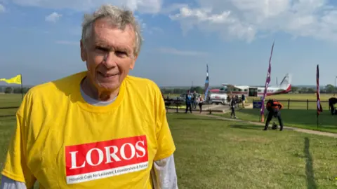 BBC David Hartridge in a yellow T-shirt with a LOROS logo standing in from of the plane he jumped out of