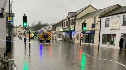 Michelle Webley A street through Lydney in Gloucestershire is seen flooded with a fire engine visible in the distance. The water is several inches deep