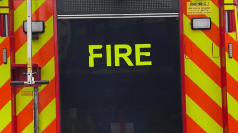 The rear of a fire engine, with yellow and orange high visibility markings and yellow text on a dark panel which reads "fire" in capital letters.