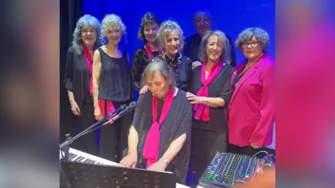 Silhouette Band Women gathered around a keyboard piano wearing black and pink and smiling as one woman plays.