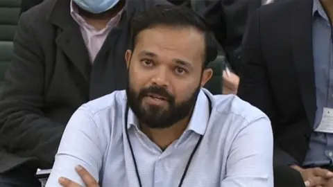 House of Commons/PA Azeem Rafiq, a bearded man at an official hearing. He is wearing a light blue shirt with a black lanyard around his neck and he looks emotional.