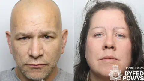 Dyfed-Powys Police Police mugshots of Geraint Berry and Michelle Mills after their arrests. He is a 46-year-old man with no hair, wearing a grey t-shirt. She is a 46-year-old woman with brown wavy hair.