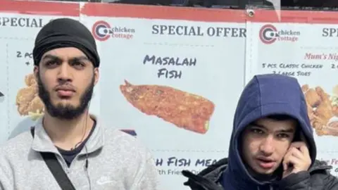 Qureshi family Abdul Aziz Ansari - a dark beard and wearing a grey Nike top and black beanie hat, standing next to Temur Qureshi, who has a blue hooded top and black coat, with his left hand touching his left ear 