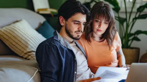 Getty Images A man and woman sitting in a front room and looking anxiously at bills 