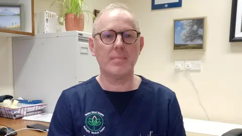 Dr John Diamond, who has thinning grey hair and glasses and who is wearing navy medical scrubs with his name on the chest, sits at a desk with a computer and some papers on it. 
