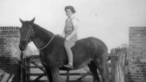 British Resistance Museum A blurry black and white photo showing Jill Monk on a horse, with a gate and wall behind her. She has short fair hair and is wearing a knitted tank top over a white shirt,  jodhpurs and lace-up shoes. The horse is dark coloured and both are looking at the camera. 