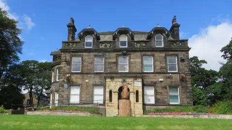 Philandju/Geograph A three-storey residence built in the 1700s and featuring Georgian bow windows looms over a sloped lawn with blue skies above and trees seen on either side. Flower beds can be seen running across the front of the property on either side of a small statue. 