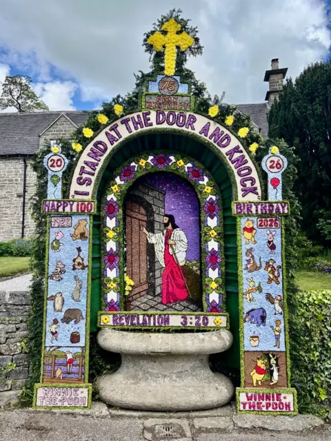 Tissington Well Dressings A large stone well surrounded by a huge floral display. The display includes an image of Jesus made from petals and natural materials. The words "I stand by the door and knock" are included in the design. On the columns of the design are images of Winnie the Pooh and other characters. There is a large yellow crucifix at the top of the display.