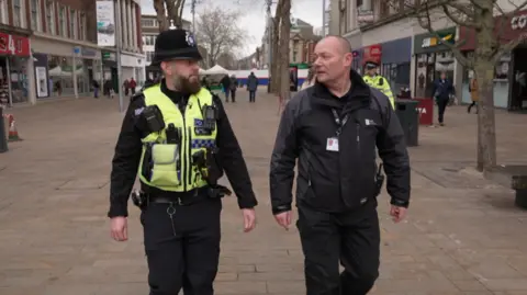 Gareth Bielby on the left and Shaun Casson on the right. Both are walking down Hull's City Centre and are talking to each other. Gareth is dressed in police uniform with a yellow high vis vest, it has a police radio and body cam attached to it. Shaun is dressed in black, including a black coat and trousers.