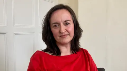 A woman with dark hair and wearing a red top sitting down in white-panelled room