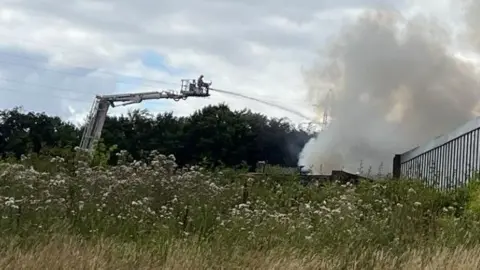 Andy Watson/BBC Firefighters on an aerial ladder platform spraying water at the fire, which is evaporating in a cloud of white smoke, contrasting the deep black of the fire itself.