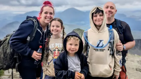 Family photo Edison has blonde curly hair and is wearing a navy jacket and grey T-shirt. He is standing next to his sister, who is smiling and has her long brown hair plaited, and also next to his brother, who has brown hair and is wearing a cream hoodie. Their dad, wearing a black T-shirt and rucksack, and mum, who has a navy hoodie and heavy rucksack, are standing next to them all. There are mountains in the background.