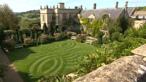 A picture shows Euridge Manor and its grounds slightly from above. The walled gardens have a green lawn in the centre cut into a butterfly shape, with an ivy-covered stately home behind. 