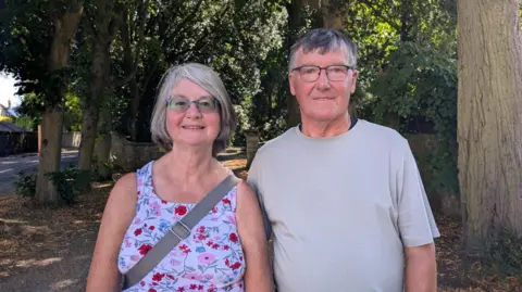 A man and a woman stand in shade under green trees with dappled sunlight breaking through the leaves. The woman has grey hair in a bob and wears blue-framed glasses and a floral print dress. The man has short grey hair and wears glasses and a light grey T-shirt.