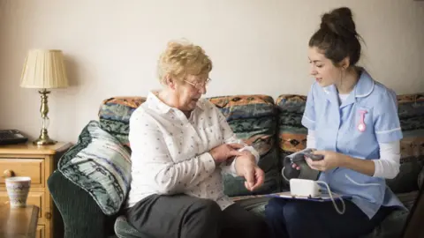 A nurse checks the blood pressure of an older lady. They are sat on a sofa in a domestic setting.