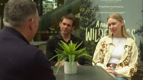Nik Payne, who has dark hair and is wearing a black open-collared shirt, and Ronja, who is blonde and wears a cream overshirt over a white top, sit in front of a poster for the film and speak to the camera