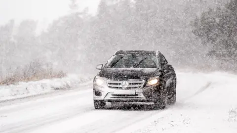 PA Media A black Mercedes Benz has its headlights and windscreen wipers on as it navigates through a snowstorm on a road which is already covered in snow in the North York Moors National Park