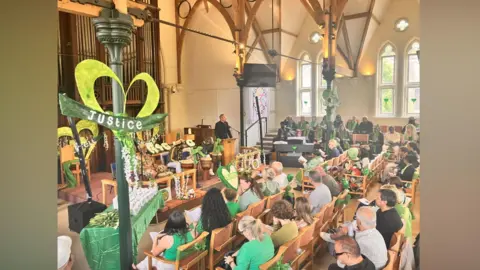 A large congregation sit in a church which is decorated with green banners.