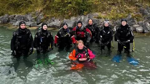 Rob Mason A group of people are stood knee-deep in outdoor water dressed in full scuba diving suits and equipment.