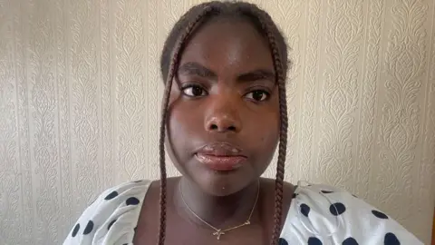 Kayla Kayla has two braids falling either side of her face. She is wearing a thin gold chain around her neck and a black and white polka dot top. 